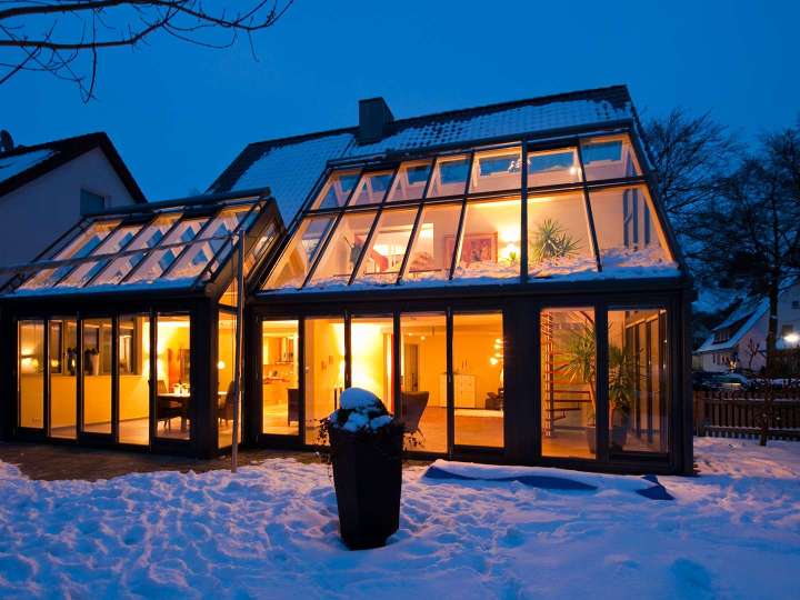 zwei beleuchtete Wintergärten an einem Wohnhaus bei Dämmerung im Schnee